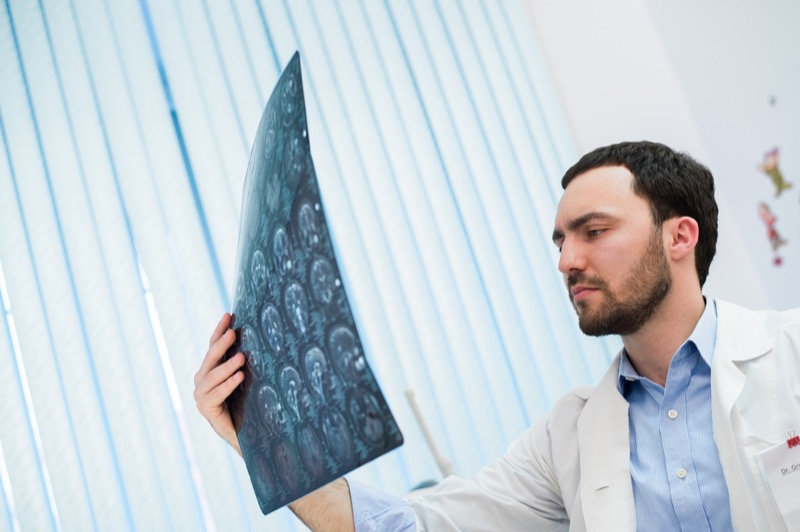 Closeup portrait of intellectual man healthcare personnel with white labcoat, looking at brain x-ray radiographic image, ct scan, mri, clinic office background. Radiology department