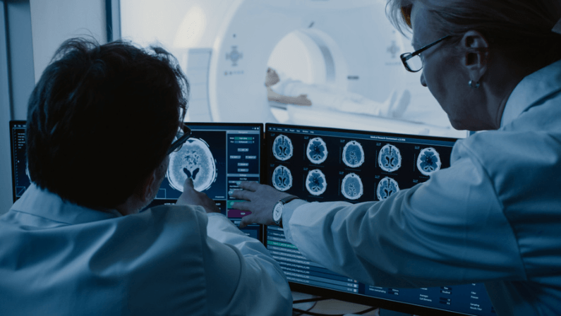 In Control Room Doctor and Radiologist Discuss Diagnosis while Watching Procedure and Monitors Showing Brain Scans Results, In the Background Patient Undergoes MRI or CT Scan Procedure.